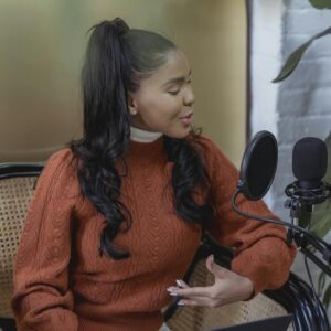 Confident young ethnic female blogger with long dark hair in casual clothes sitting on chair with laptop and speaking into microphone while recording podcast at home