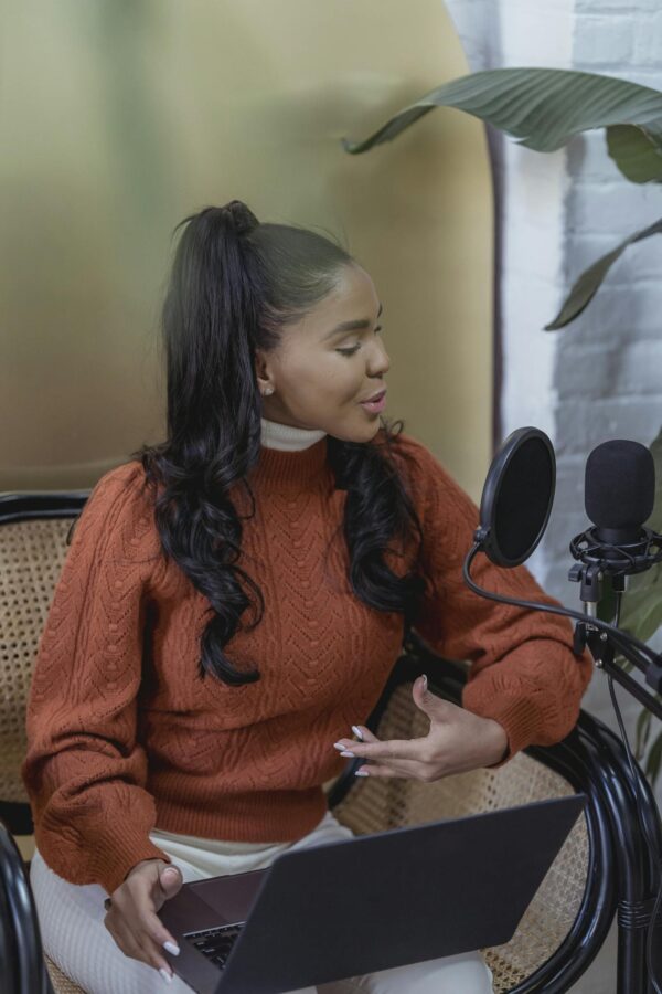 Confident young ethnic female blogger with long dark hair in casual clothes sitting on chair with laptop and speaking into microphone while recording podcast at home