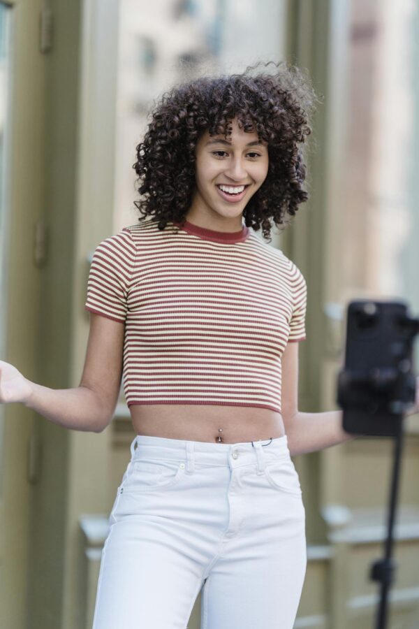 Positive ethnic female in stylish outfit recording live broadcast on smartphone while standing on street with buildings on blurred background