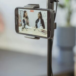 Young diverse women sitting at table with microphones and gadgets and talking while recording podcast on smartphone placed on tripod in studio