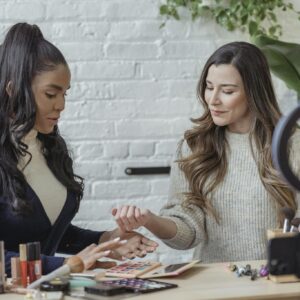 Young positive multiethnic female bloggers testing makeup products while sitting at table and recording vlog on smartphone placed on tripod with ring light